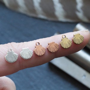 May include: Five small seashell charms in different metal finishes: silver, rose gold, gold, and brass. The charms are shown on a finger.