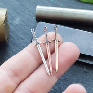 May include: Three miniature sword charms in silver, rose gold, and black metal. The charms are held in a hand against a gray background.