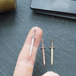 May include: Three miniature sword charms in silver, copper, and gold finishes. The charms are shown on a finger for scale.