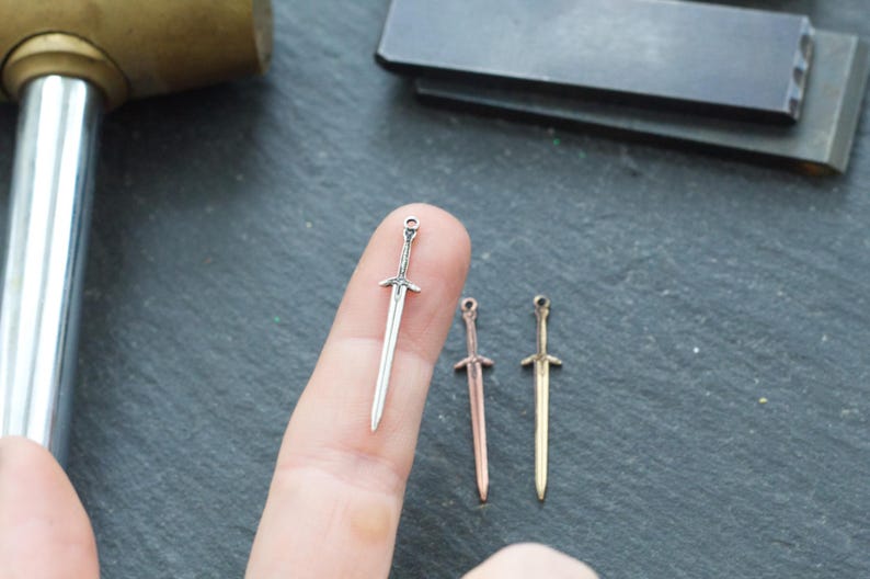 May include: Three miniature sword charms in silver, copper, and gold finishes. The charms are shown on a finger for scale.