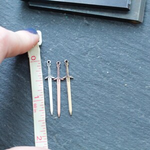 May include: Three silver sword charms, each about 1 inch long, are lined up next to a ruler. The swords are different shades of silver, with one being a rose gold color.
