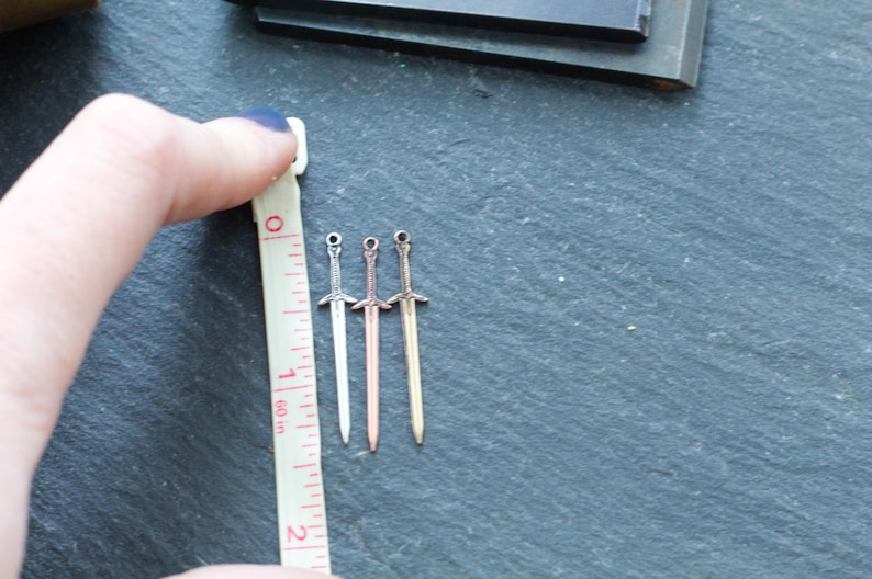May include: Three silver sword charms, each about 1 inch long, are lined up next to a ruler. The swords are different shades of silver, with one being a rose gold color.