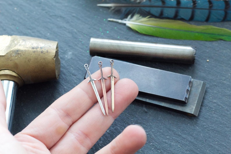 May include: Three miniature sword charms in silver, rose gold, and black metal. The charms are held in a hand against a gray background.