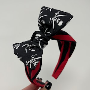Puede incluir: Una diadema negra con un lazo con un estampado de calaveras y huesos cruzados. La diadema tiene rayas rojas y negras. Este accesorio de temática pirata presenta un lazo central para un toque decorativo.