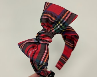 May include: A headband featuring a large, knotted bow on top, covered in a red, green, blue, and yellow plaid fabric pattern. The fabric has a smooth texture and the bow is positioned centrally on the band, which is also wrapped in the same plaid material.