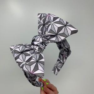 May include: A gray and white headband with a large bow. The bow is made of fabric with a geometric pattern of stars.