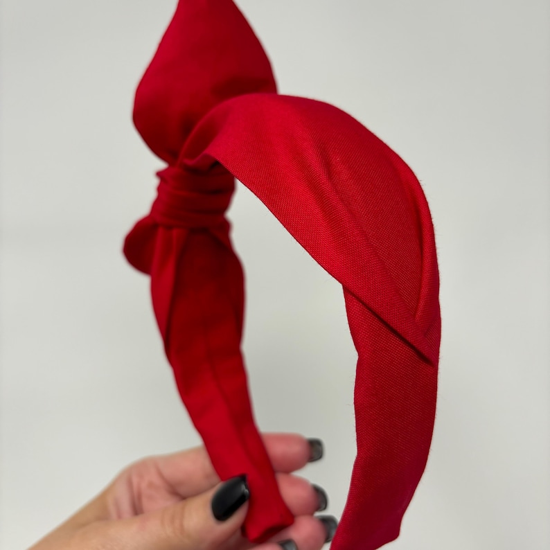 May include: A red fabric headband with a knotted bow detail. The headband is made of a soft, stretchy material and is designed to be comfortable to wear.