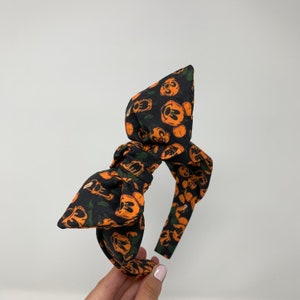 May include: A black headband with a large orange and black bow. The bow is made of fabric with a pattern of orange Mickey Mouse heads on a black background.