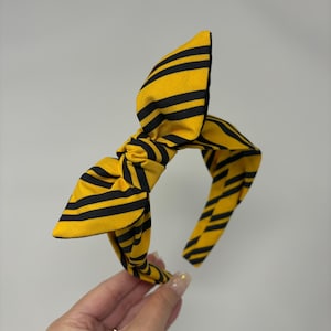 May include: A yellow and black striped headband with a large bow. The headband is made of fabric and has a simple design.