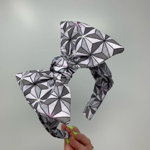 May include: A gray and white headband with a large bow. The bow is made of fabric with a geometric pattern of stars.
