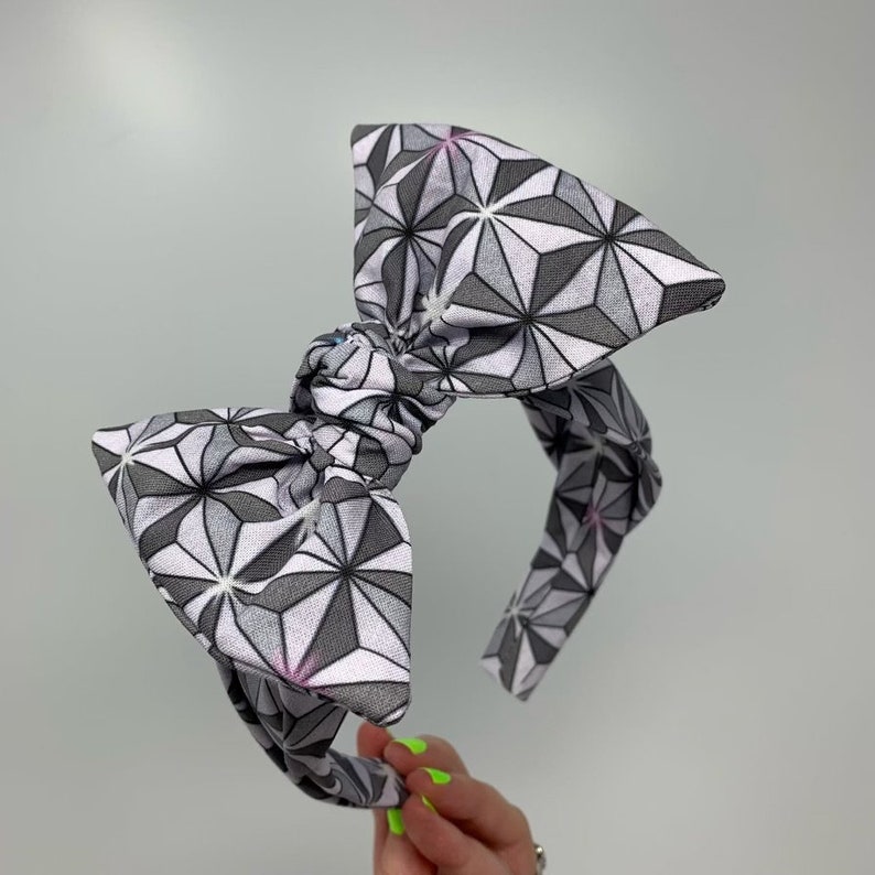May include: A gray and white headband with a large bow. The bow is made of fabric with a geometric pattern of stars.