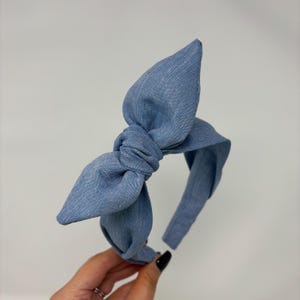 May include: A light blue denim headband with a large bow. The bow is knotted in the center and has two large loops. The headband is a solid color and is made of a soft fabric. This hair accessory is perfect for adding a touch of style.