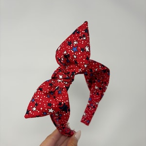 May include: Red fabric headband with a bow, featuring a pattern of white dots, white stars, and blue stars. The headband is a festive accessory, perfect for holidays or themed events.