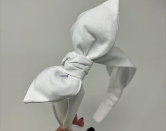 May include: A solid white headband featuring a large, structured fabric bow centered on the top. The bow is tied in a knot style with two distinct loops extending outward, and the headband band is covered in the same smooth, plain white fabric.