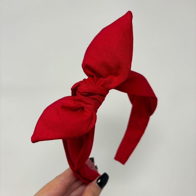May include: A red fabric headband with a large bow on top. The headband is made of a soft, stretchy material and is perfect for adding a touch of style to any outfit.