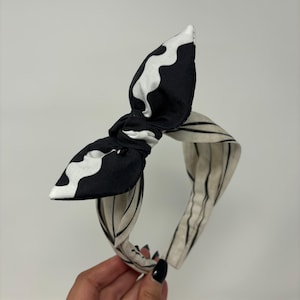 May include: A black and white patterned headband with a knotted bow. The headband is made of fabric and has a striped pattern.