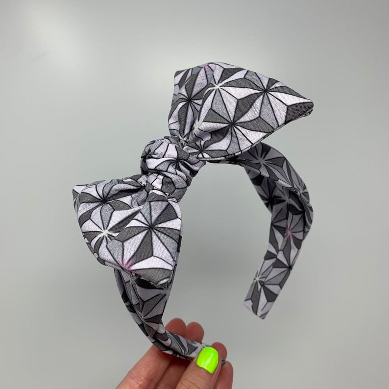 May include: A gray and white patterned headband with a large bow. The headband is made of fabric and has a geometric design.