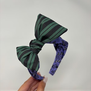 Könnte beinhalten: Stoff-Haarreif mit großer Schleife. Die Schleife hat grün-schwarze Streifen, während das Stirnband selbst lila ist und ein Muster aus Noten und kleinen Illustrationen aufweist. Ein Haar-Accessoire.