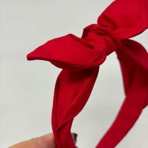 May include: A red fabric headband with a large bow. The headband is twisted and knotted to create the bow.