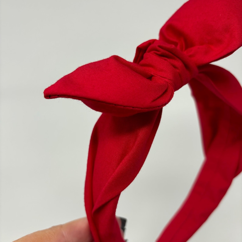 May include: A red fabric headband with a large bow. The headband is twisted and knotted to create the bow.