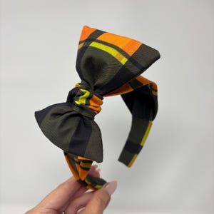 Può includere: Una fascia a quadri con un grande fiocco. Il fiocco e la fascia presentano un motivo arancione, giallo e nero. Un accessorio di moda.