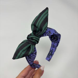 Könnte beinhalten: Ein Stoff-Haarreif mit einer Schleife. Die Schleife hat grün-schwarze Streifen, während das Stirnband lila mit einem schwarz-weißen Muster ist. Das Stirnband wird von einer Hand gehalten.
