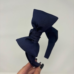 May include: A navy blue headband with a large bow. The headband is made of a soft, fabric material.