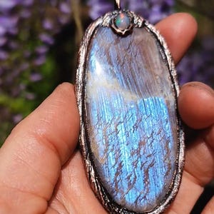 Puede incluir: Colgante ovalado con una gran piedra preciosa azul iridiscente con superficie texturizada. La piedra está engastada en un marco de metal color cobre, coronada por una pequeña piedra opalescente redonda. Fondo borroso con flores moradas.