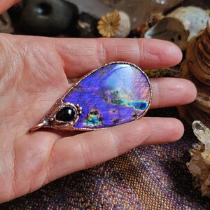 May include: A teardrop-shaped pendant made of copper wire and a purple labradorite gemstone. The pendant features a small black gemstone accent.