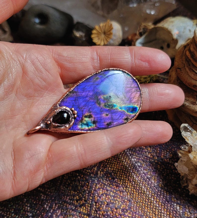 May include: A teardrop-shaped pendant made of copper wire and a purple labradorite gemstone. The pendant features a small black gemstone accent.