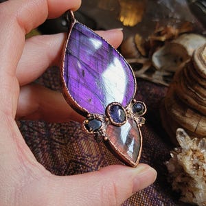 May include: A copper wire wrapped pendant featuring a large purple labradorite gemstone with iridescent flashes. The pendant is accented with smaller amethyst and black stones.