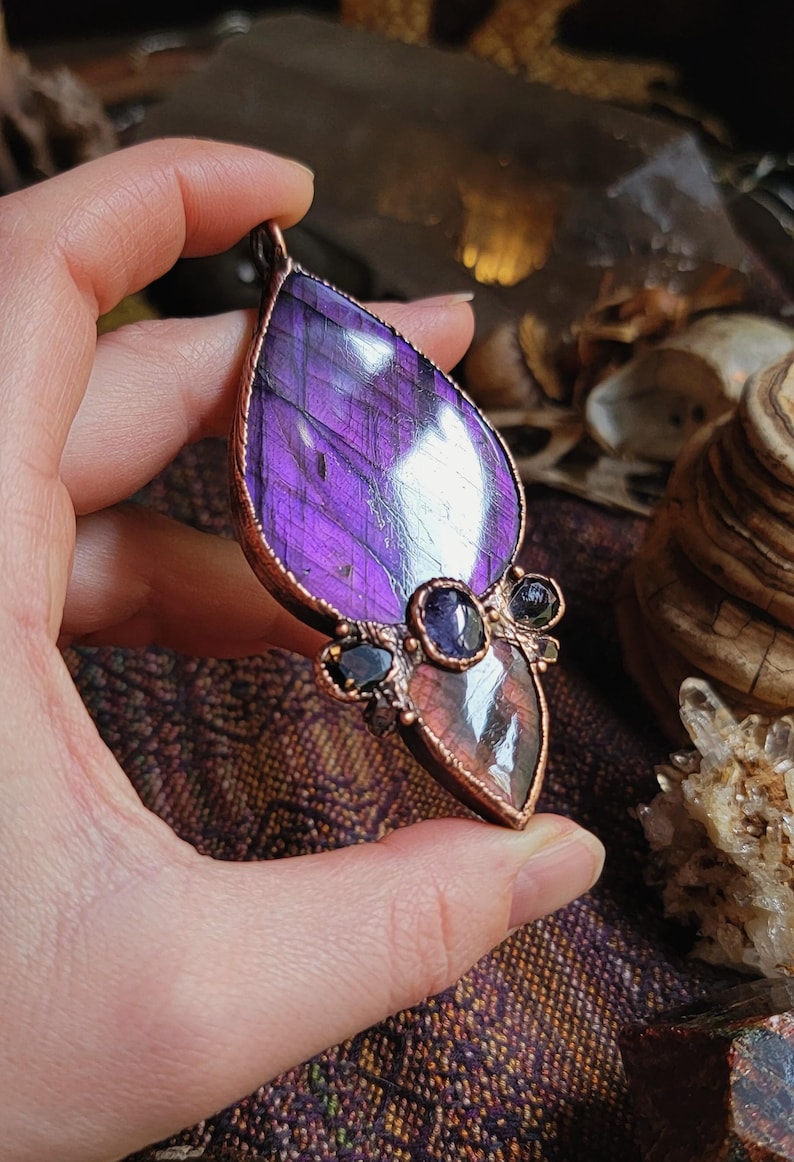 May include: A copper wire wrapped pendant featuring a large purple labradorite gemstone with iridescent flashes. The pendant is accented with smaller amethyst and black stones.