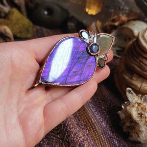 May include: A copper wire wrapped pendant featuring a large purple labradorite stone with iridescent shimmer. The pendant is accented with smaller stones in shades of blue and brown.