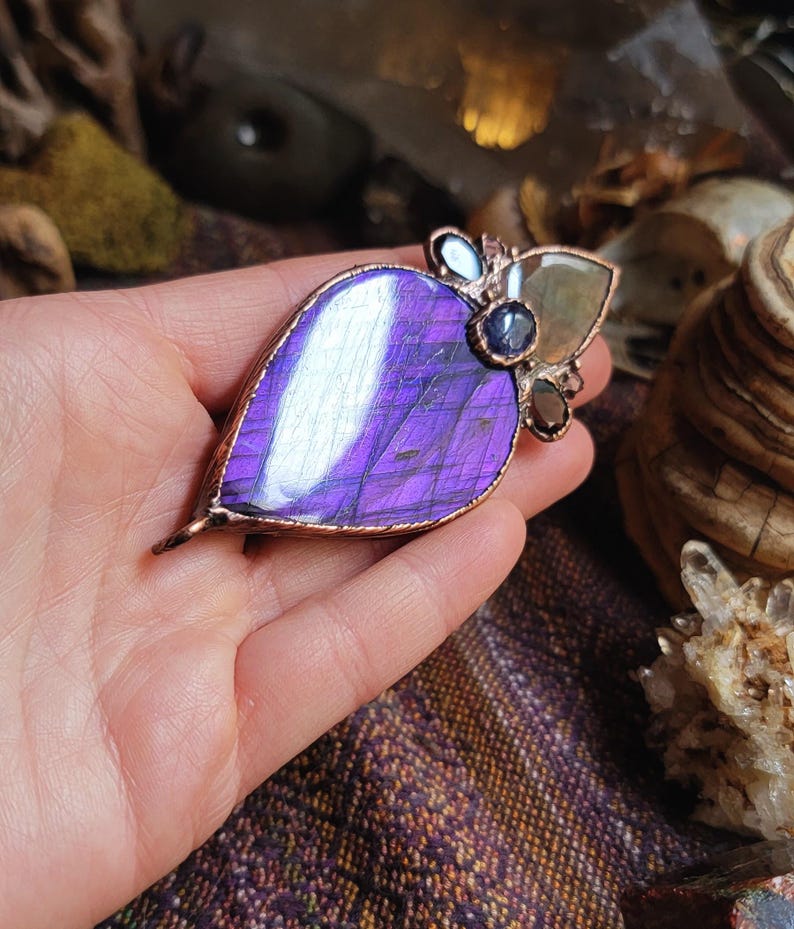 May include: A copper wire wrapped pendant featuring a large purple labradorite stone with iridescent shimmer. The pendant is accented with smaller stones in shades of blue and brown.
