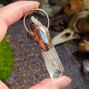 May include: A clear, pointed crystal pendant encased in a textured, oxidised copper cap. The cap features a small, smooth, light blue oval inlay and is attached to a simple, round copper wire bail at the top for hanging as a necklace charm.