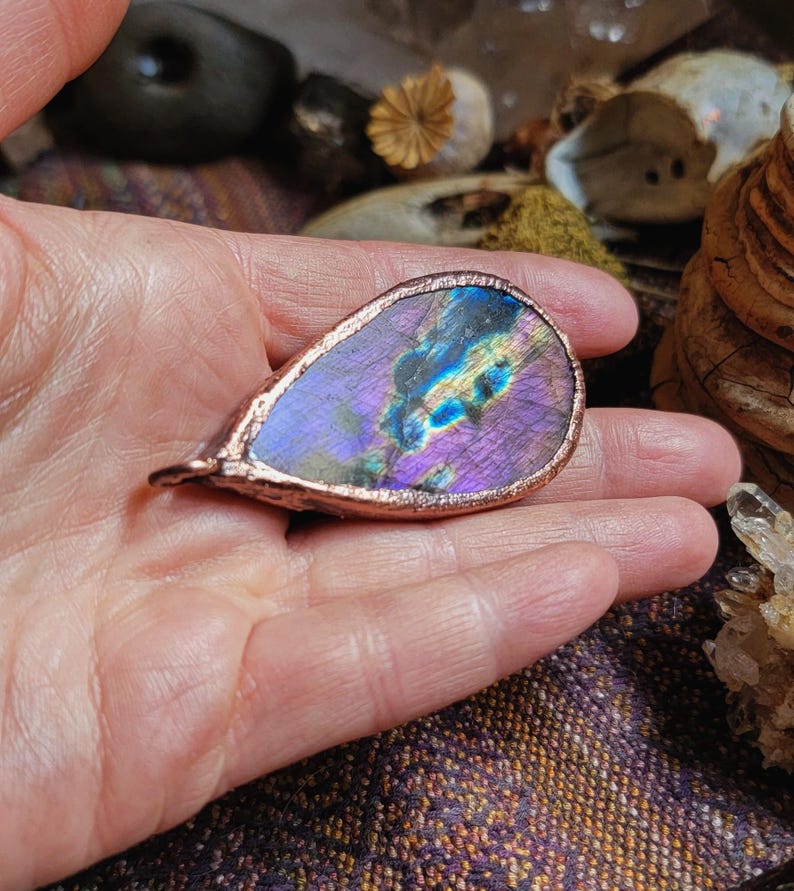 May include: A teardrop-shaped pendant with a copper frame and a iridescent labradorite stone. The stone has a purple, blue, and green color.