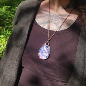 May include: A teardrop-shaped pendant necklace with a copper chain. The pendant is made of iridescent glass with a dark purple gemstone at the top.