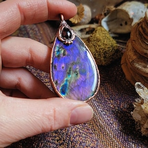 May include: A teardrop-shaped pendant with a copper wire frame and a purple labradorite stone. The stone has a iridescent sheen and a dark purple garnet accent at the top.