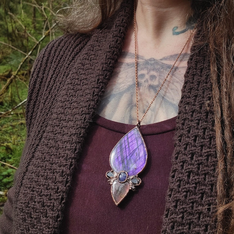 May include: A purple and iridescent labradorite gemstone pendant with copper wire accents and smaller blue gemstones. The pendant is hanging from a copper chain necklace.