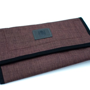 May include: A brown fabric pouch with a black trim and a small leather patch with a logo. The pouch is folded in half and is lying on a white surface.