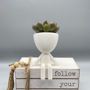 May include: White ceramic planter shaped like a person sitting with legs crossed, holding a small succulent plant. The planter is sitting on a stack of white books with the text "follow your dreams" printed on the front.