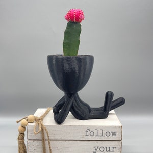 May include: A black ceramic planter shaped like a person laying down with a pink cactus plant inside. The planter is sitting on a stack of white books with the words "follow your dreams" printed on the front.