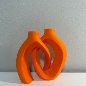 May include: A pair of orange ceramic candle holders with a modern abstract design. The holders are intertwined and have a smooth, glossy finish.