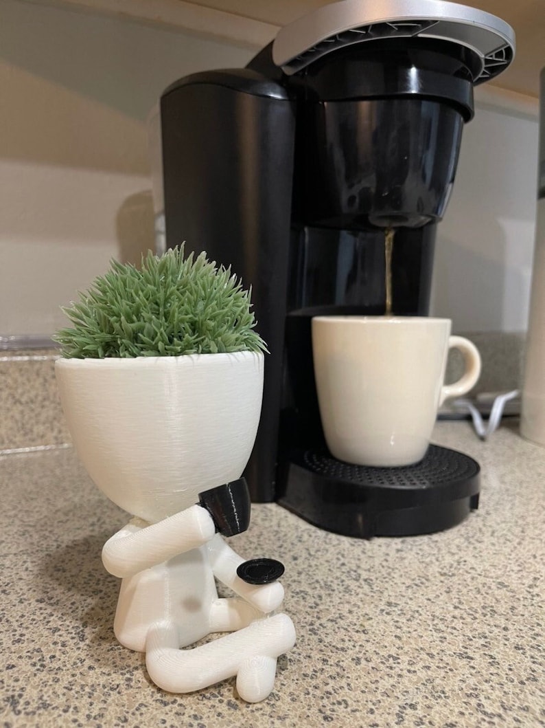 May include: A white 3D-printed planter shaped like a person, holding a small black saucer. The planter holds a green, textured plant. A black coffee maker and a white mug are in the background.