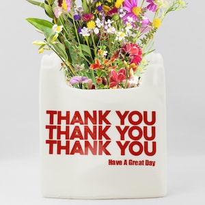 May include: White ceramic vase shaped like a grocery bag with red text that says "Thank You" three times and "Have A Great Day". The vase is filled with a bouquet of colorful flowers.