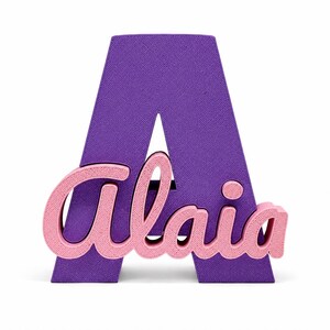 May include: A large, purple, three-dimensional letter A with a textured surface. Attached to the front of the letter is a smaller, light pink, three-dimensional script nameplate that reads Alaia in a cursive font with a matching textured finish.