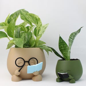 May include: Two whimsical plant pots, one tan and one green, each with a face and holding a book. The tan pot has black glasses and a blue book, while the green pot has a black book. Both pots contain green plants with varying leaf patterns.