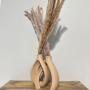 May include: Two beige ceramic vases with a unique, abstract design. The vases are filled with dried pampas grass.
