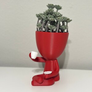 May include: A red figurine planter with a head filled with green plants, holding a white teacup and saucer. The figurine is sitting down, with its legs crossed. The planter is a unique decorative item.