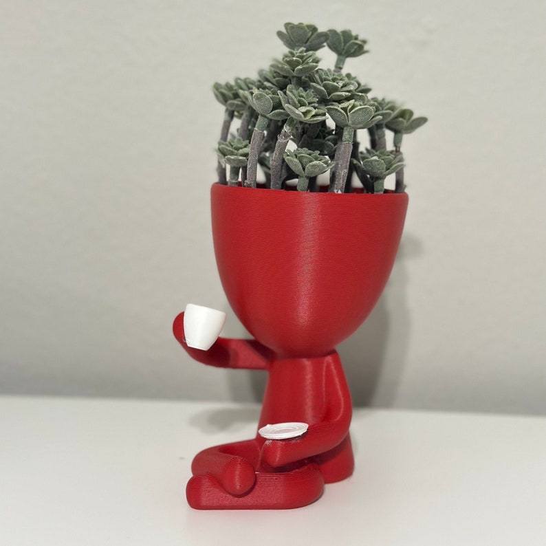 May include: A red figurine planter with a head filled with green plants, holding a white teacup and saucer. The figurine is sitting down, with its legs crossed. The planter is a unique decorative item.